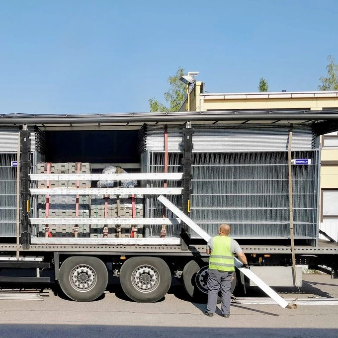Työntekijä huomioliivissä purkaa pitkiä metallipalkkeja kuorma-autosta, joka kuljettaa metalliaitoja ja betonilohkoja.