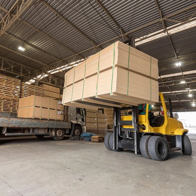 Keltainen trukki nostaa isoa vanerilevyjen kasaa varastossa, taustalla näkyy lisää vanerikasoja ja kuorma-auto.